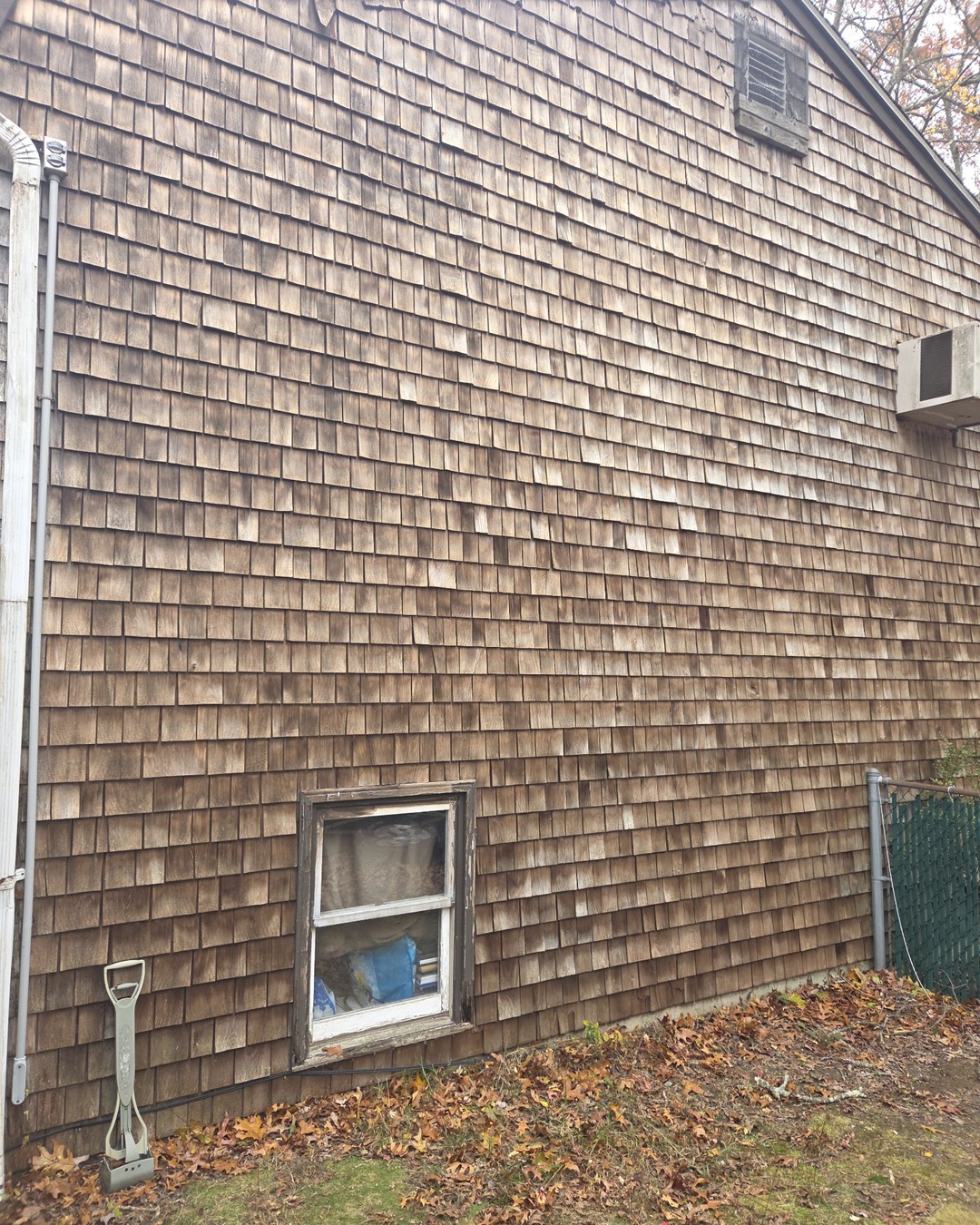 Weathered brown cedar shake siding on a side gable wall with an unpainted basement window frame before exterior painting