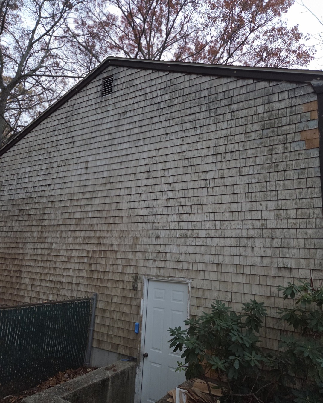 Gray-brown wood shingle gable with a white walkout basement door and newly replaced bare wood shingles in the upper corner before painting