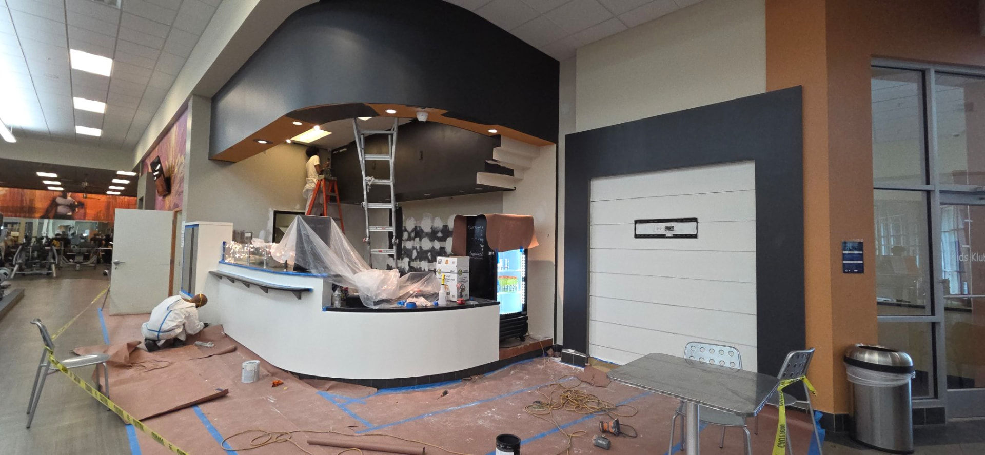 Commercial gym reception area mid-painting with a painter on a ladder and floor protection in place